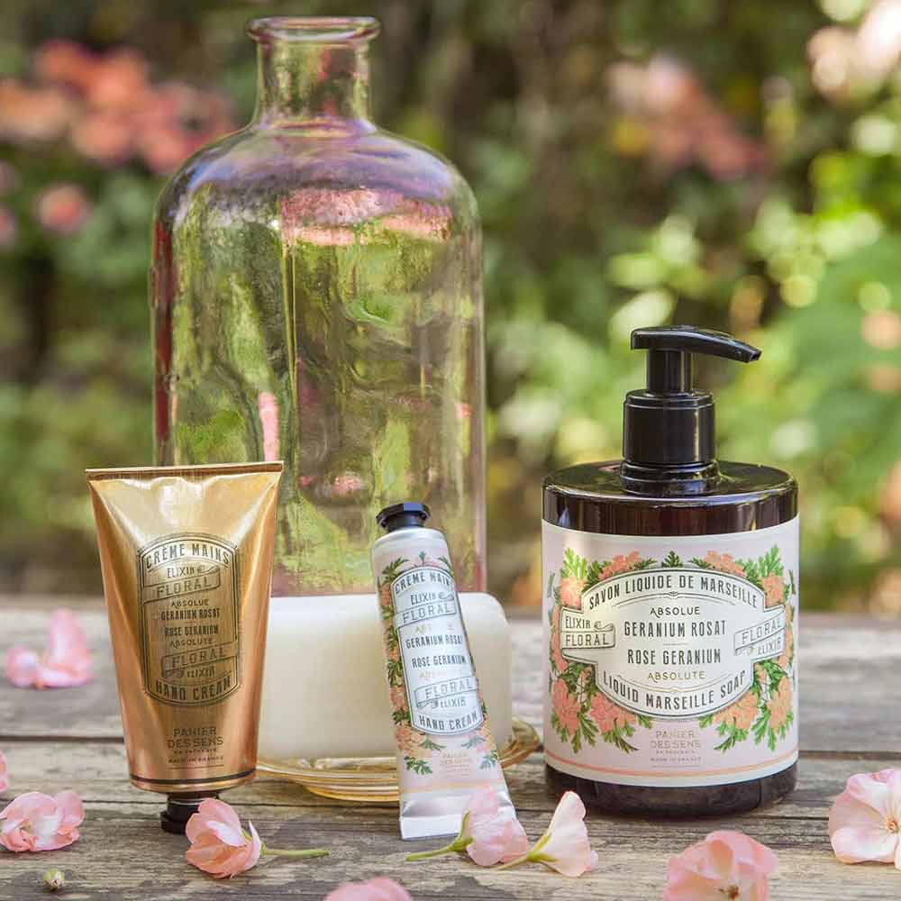 Eine Sammlung von Panier des Sens – Handcreme Rosengeranie, darunter Cremes und Flüssigseife, ausgestellt zwischen gefallenen Blütenblättern mit einer Vintage-Glasflasche im Hintergrund.