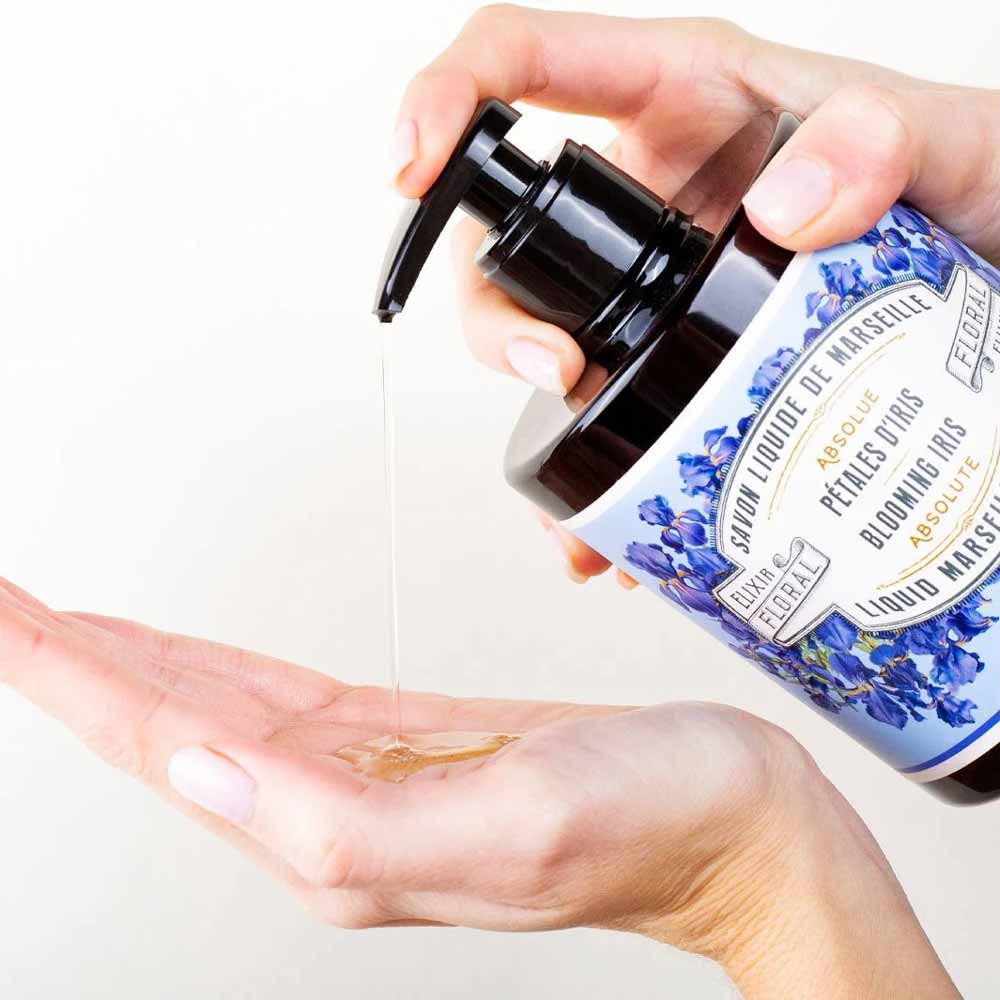 Eine Person drückt auf die Pumpe einer Flasche mit der Aufschrift „Panier des Sens – Marseiller Flüssige Handseife Irisblütenblätter 500 ml“, um Seife in ihre Hand zu geben. Die Flasche ist mit blauen Blumenmustern und einem Text versehen.