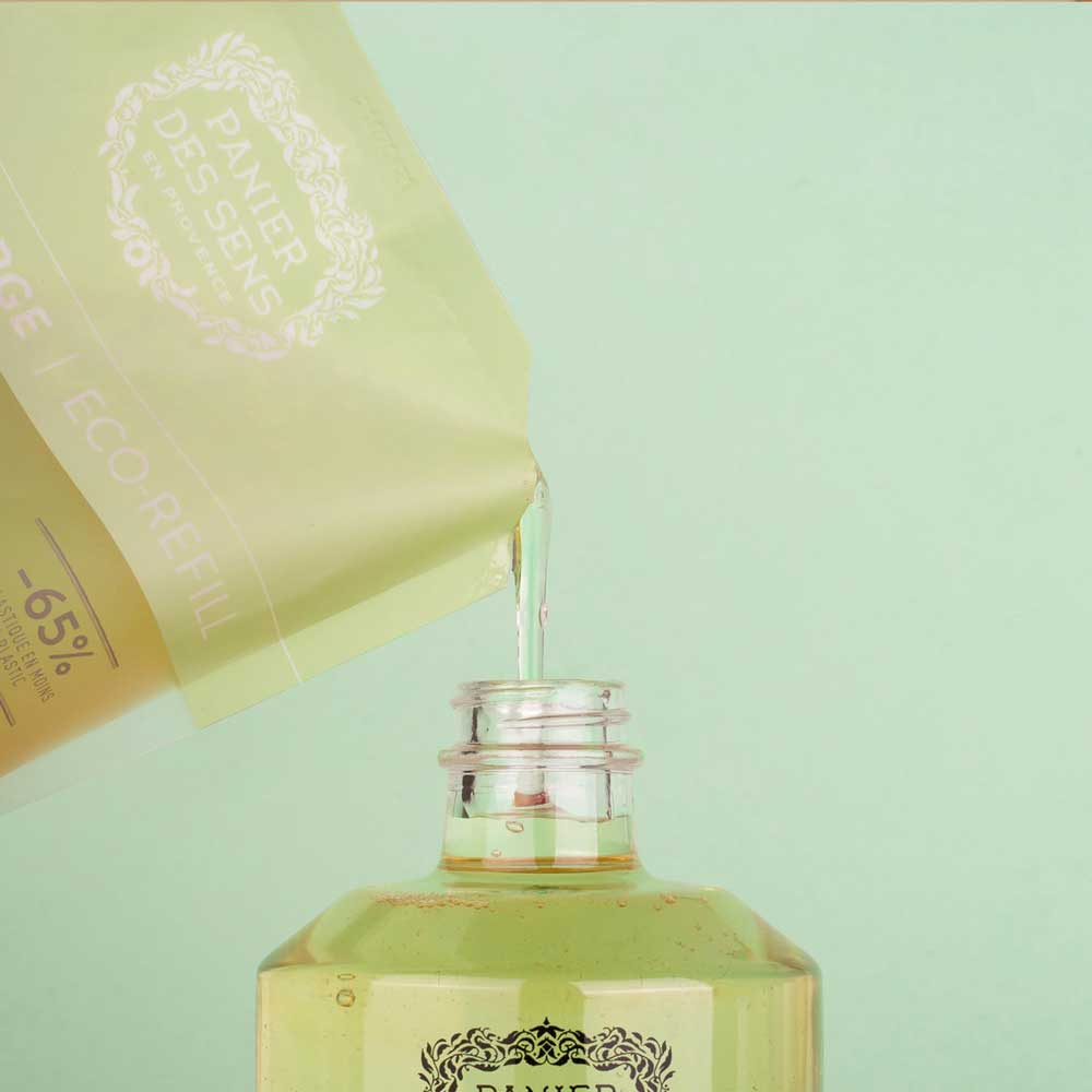 Panier des Sens - Marseiller Flüssigseife Nachfüllbeutel Jasmin 500 ml, gegossen aus einer Packung in eine Glasflasche vor einem blaugrünen Hintergrund.