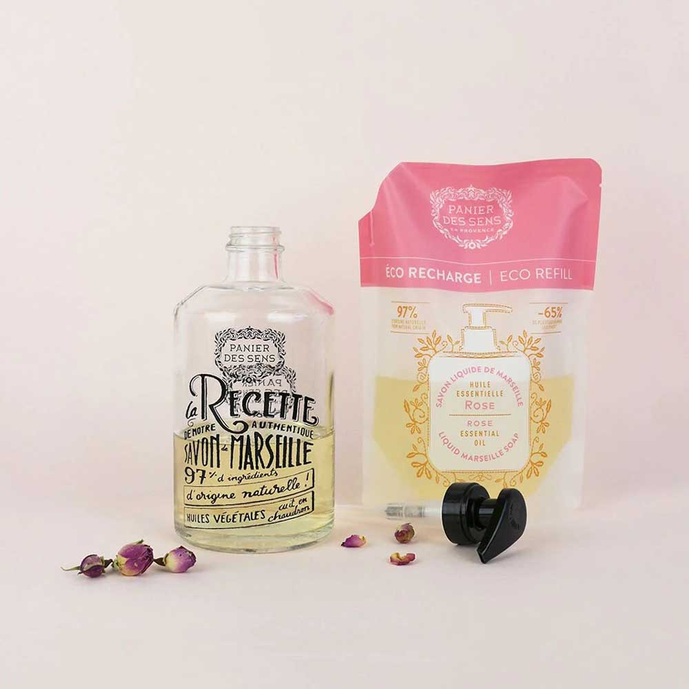 Produktpräsentation eines Panier des Sens - Marseiller Flüssigseife Nachfüllbeutel Rose 500ml und einer Öko-Nachfüllpackung mit verstreuten getrockneten Blütenblättern.