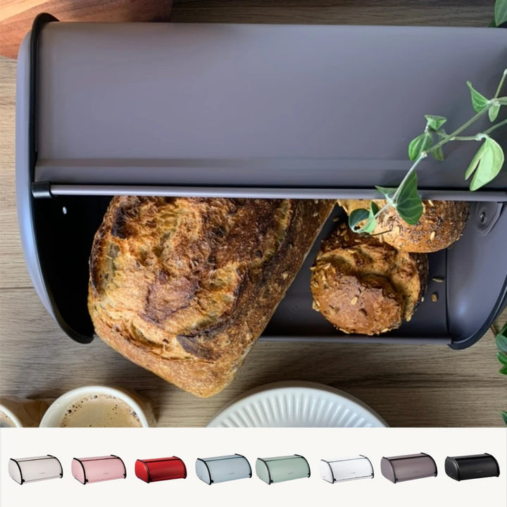 Der Plint - Brotkasten Retro Vintage, hier in acht Farben abgebildet, fasst einen Laib rustikales Brot und zwei Brötchen. Er steht matt auf einer Holzfläche neben einer Pflanze.