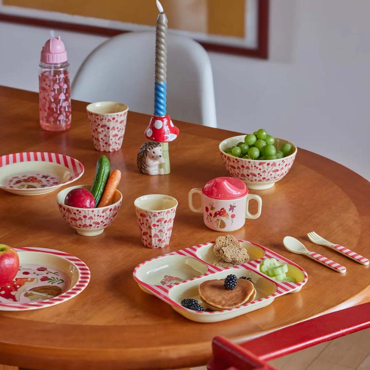 Ein Holztischset mit farbenfrohem Kindergeschirr, darunter Teller, Tassen, Schüsseln, Besteck, ein Rice - Baby Cup Forest Pink, eine Babyflasche und eine Kerze, auf dem Snacks wie Pfannkuchen, Obst und Gemüse präsentiert werden.