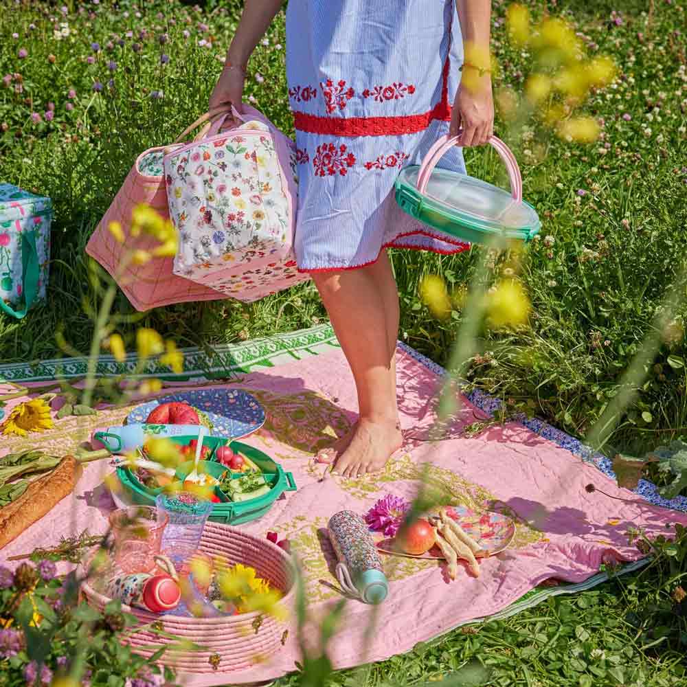 Person bereitet ein Picknick auf einer Wiese vor. Dazu gibt es eine rosa Decke, verschiedene Lebensmittel und die Reis-Kühltasche Floras Dream. Im Vordergrund sind gelbe Blumen zu sehen.