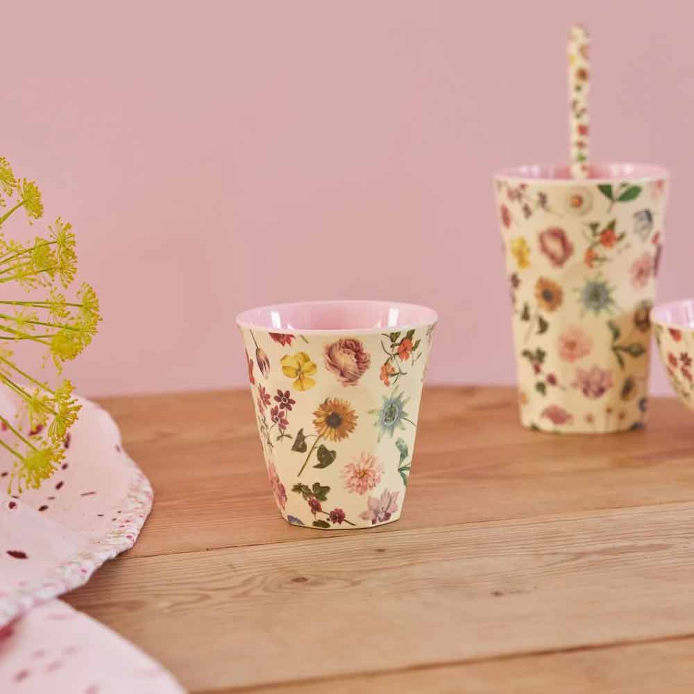 Zwei Rice - Melaminbecher Floras Dream Medium, einer davon mit Strohhalm, stehen auf einem Holztisch vor rosa Hintergrund, neben einem kleinen Blumenarrangement.