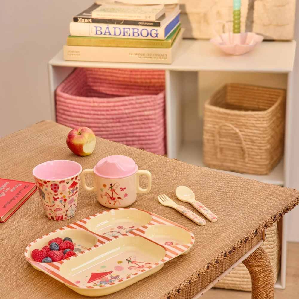 Auf einem geflochtenen Tisch steht ein Kinderteller mit Obst, Besteck und einem Rice Melaminbecher Fun Fair Medium. Im Hintergrund stehen Körbe und Bücher in den Regalen, und auf dem Tisch liegt ein Apfel.
