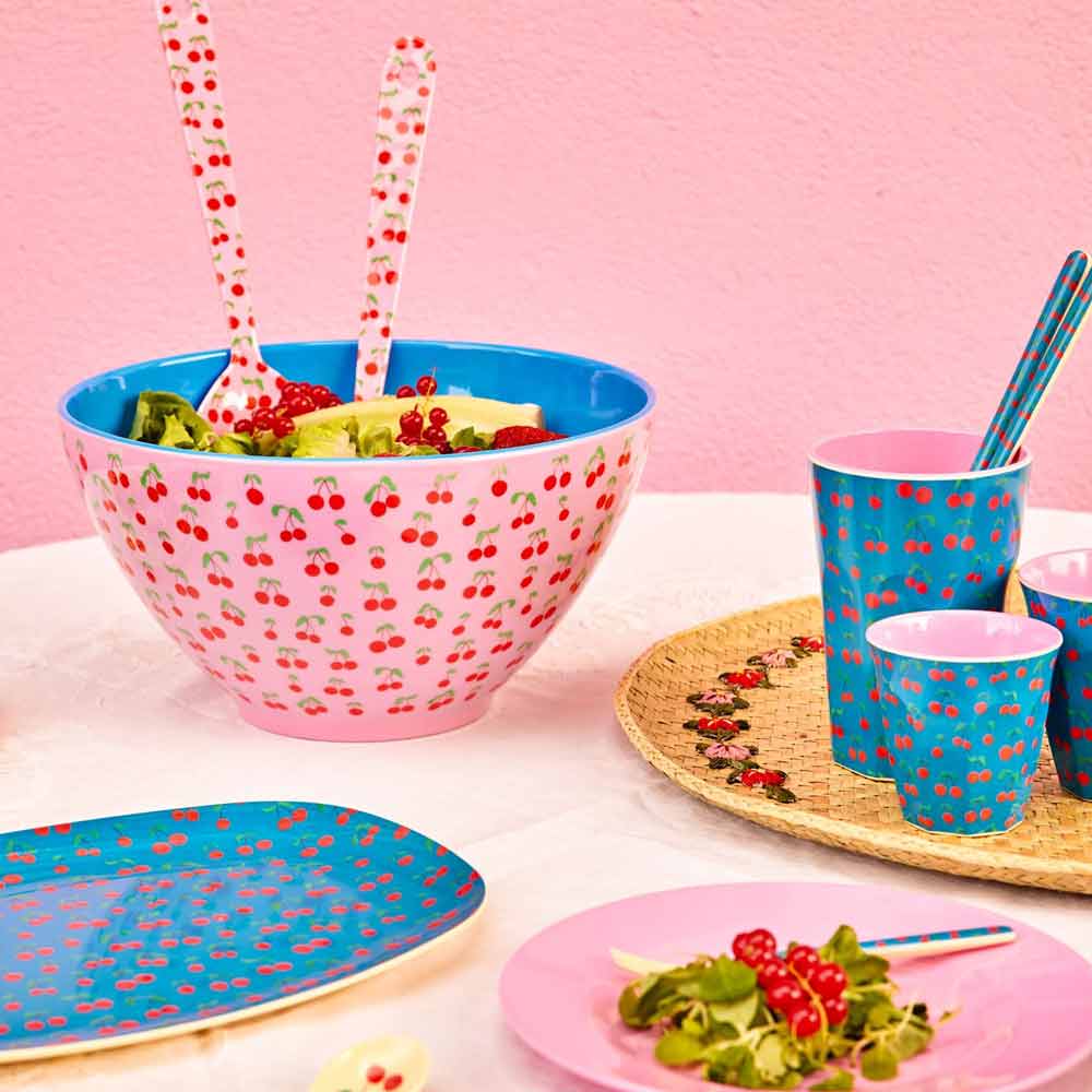 Das Rice Melamin Salatbesteck „Cherries“ besteht aus Schüsseln, Tassen und Besteck mit Kirschmuster sowie Salat in einer großen Schüssel und ist auf einem Tisch vor einer rosa Wand angeordnet.