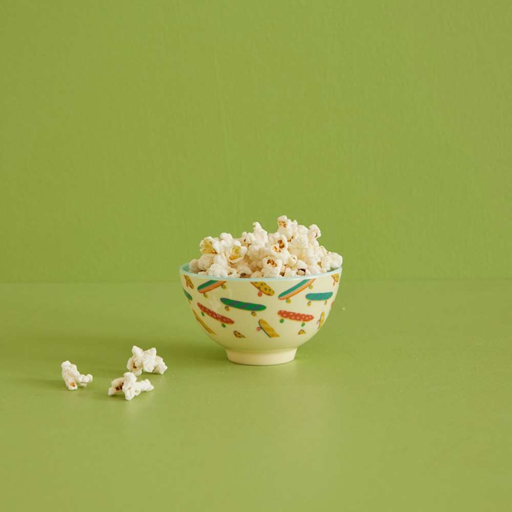 Ein kleines Rice - Melamin Schüssel Skateboard mit buntem Muster, gefüllt mit Popcorn auf grünem Untergrund, daneben sind ein paar Popcornkörner verstreut.