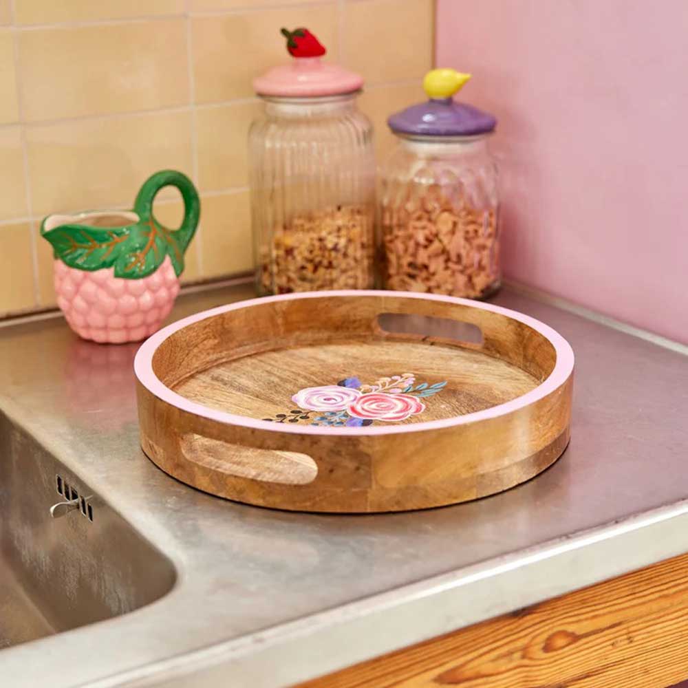 Ein rundes Holztablett von Rice mit handgemalter rosa Kante und Blumen steht auf einer Küchenarbeitsplatte. Zwei Glasgefäße mit unterschiedlichem Inhalt und ein grüner Krug stehen hinter dem Tablett in der Nähe einer gefliesten Küchenrückwand.