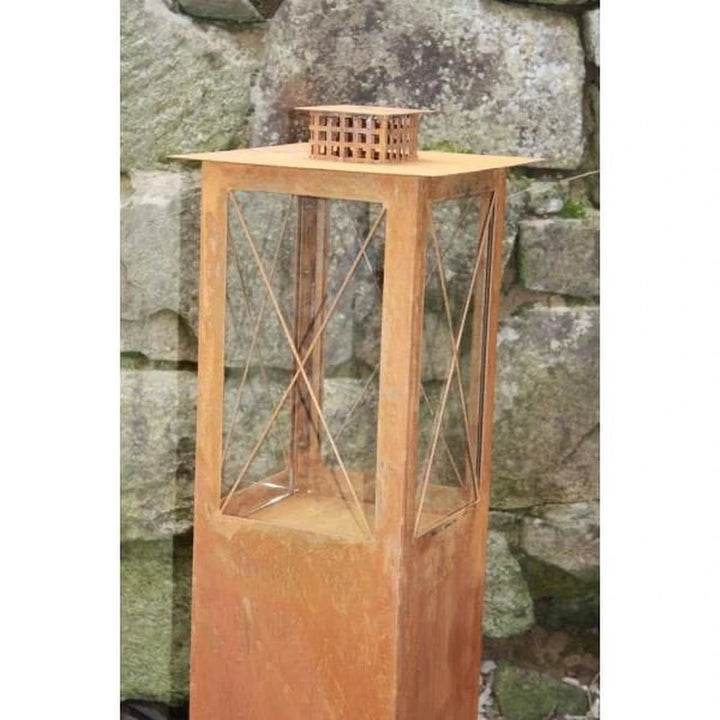 Die Rostikal Edelrost Metall Laterne Windlaterne Mega 105 cm mit Glaspaneelen und einem Rost-Finish steht vor einem Steinmauer-Hintergrund.