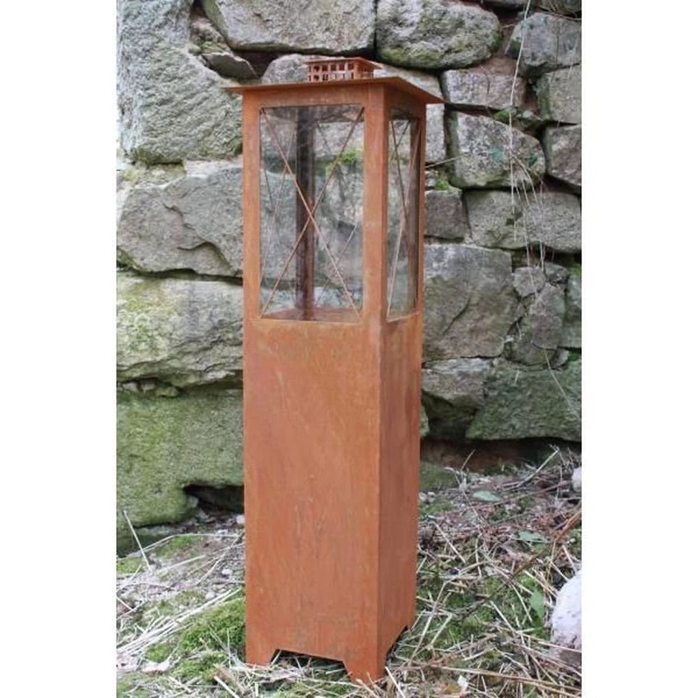 Die Rostikal Edelrost Metall Laterne Windlaterne Mega 105 cm, eine hohe rostfarbene Metalllaterne mit Glasscheiben, steht im Freien auf Gras vor einer Steinmauer.