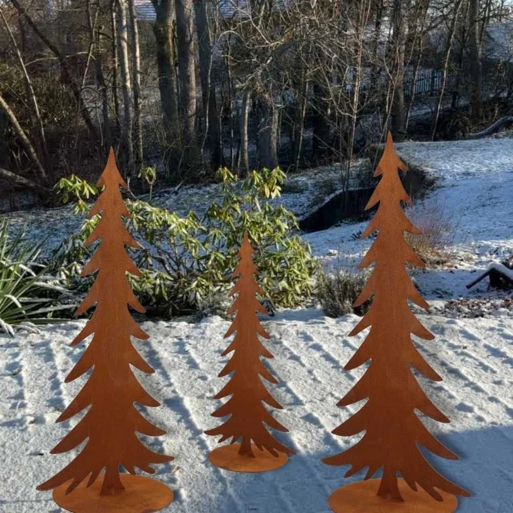 Drei Rotikal - Rost Deko Tannenbaum Skulpturen aus Metall stehen auf schneebedecktem Boden, mit echten Bäumen und Sträuchern im Hintergrund.