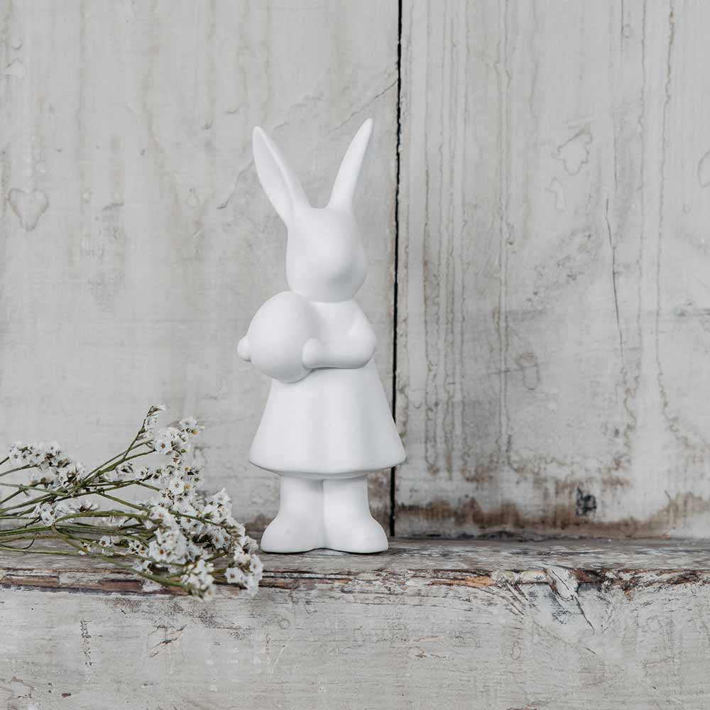 Eine weiße Figur von Storefactory – Alice Hase, die einen Hasen zeigt, der aufrecht in einem Kleid vor einem rustikalen Holzhintergrund steht, begleitet von einer kleinen Gruppe weißer Blumen.