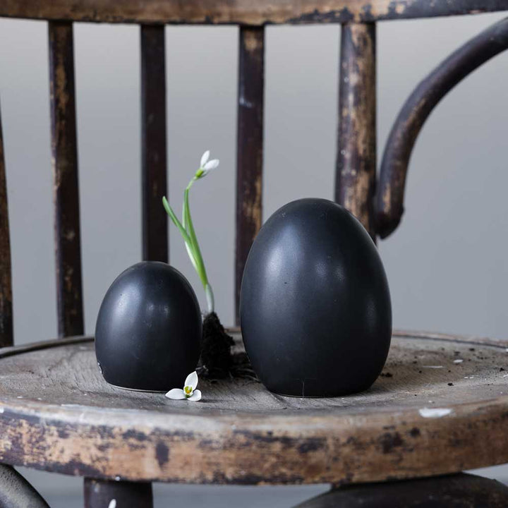 Zwei Storefactory - Bjuv Ei Keramik in verschiedenen Größen auf einem Holzstuhl mit einer Schneeglöckchenblume.