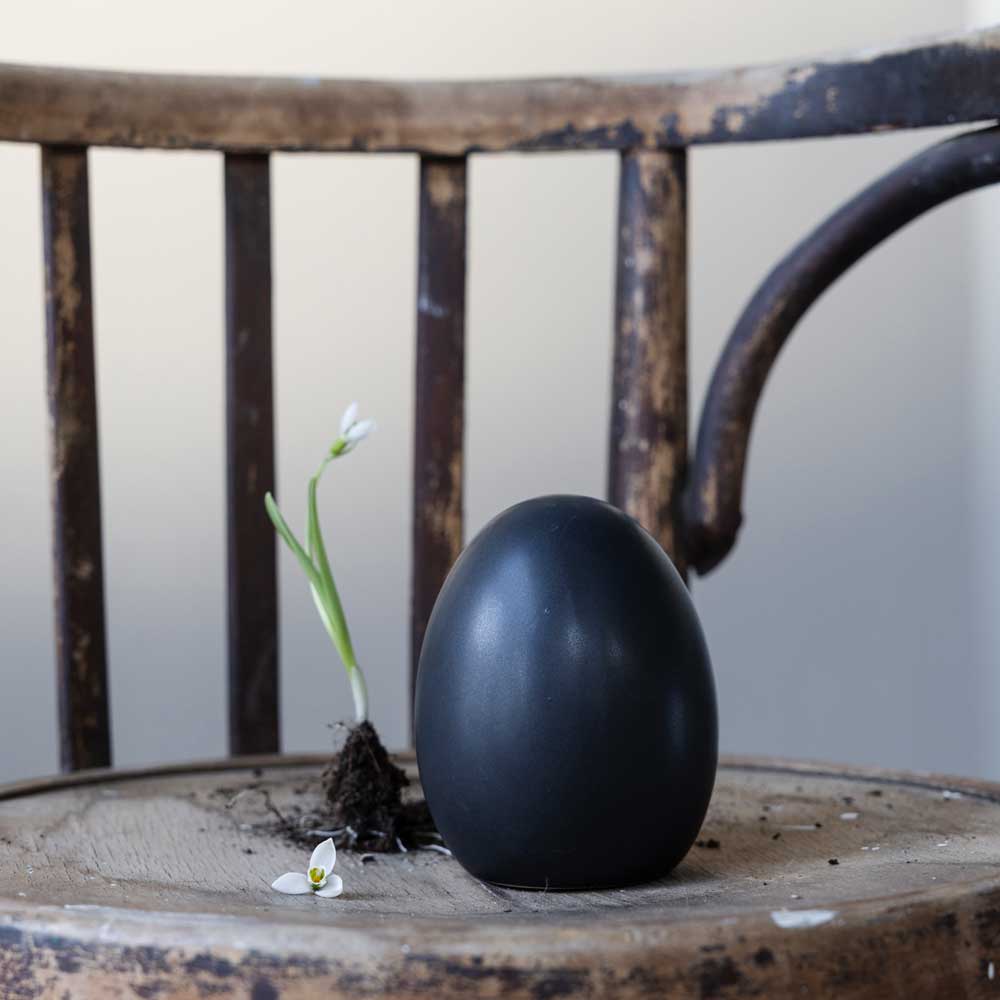 Eine Storefactory – Bjuv Ei Keramik und eine kleine weiße Blume mit ihrer Zwiebel und einer Prise Erde auf einem alten Holzstuhl.