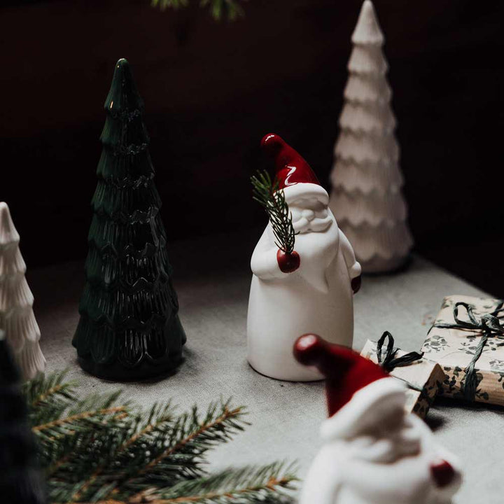 Eine festliche Weihnachtsszene mit den Bosse Weihnachtsmann-Keramikfiguren des Herstellers Storefactory, grünem und weißem Christbaumschmuck und einem verpackten Geschenk auf einem mit Tannenzweigen geschmückten Tisch.