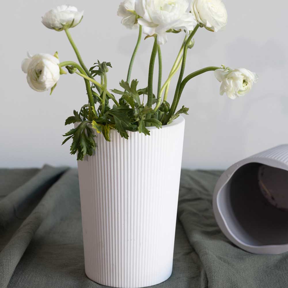 Weiße Blumen sind in einer aufrecht stehenden, gerippten Storefactory Eksberg-Vase auf einem Tisch arrangiert, neben einer passenden umgekippten Vase.