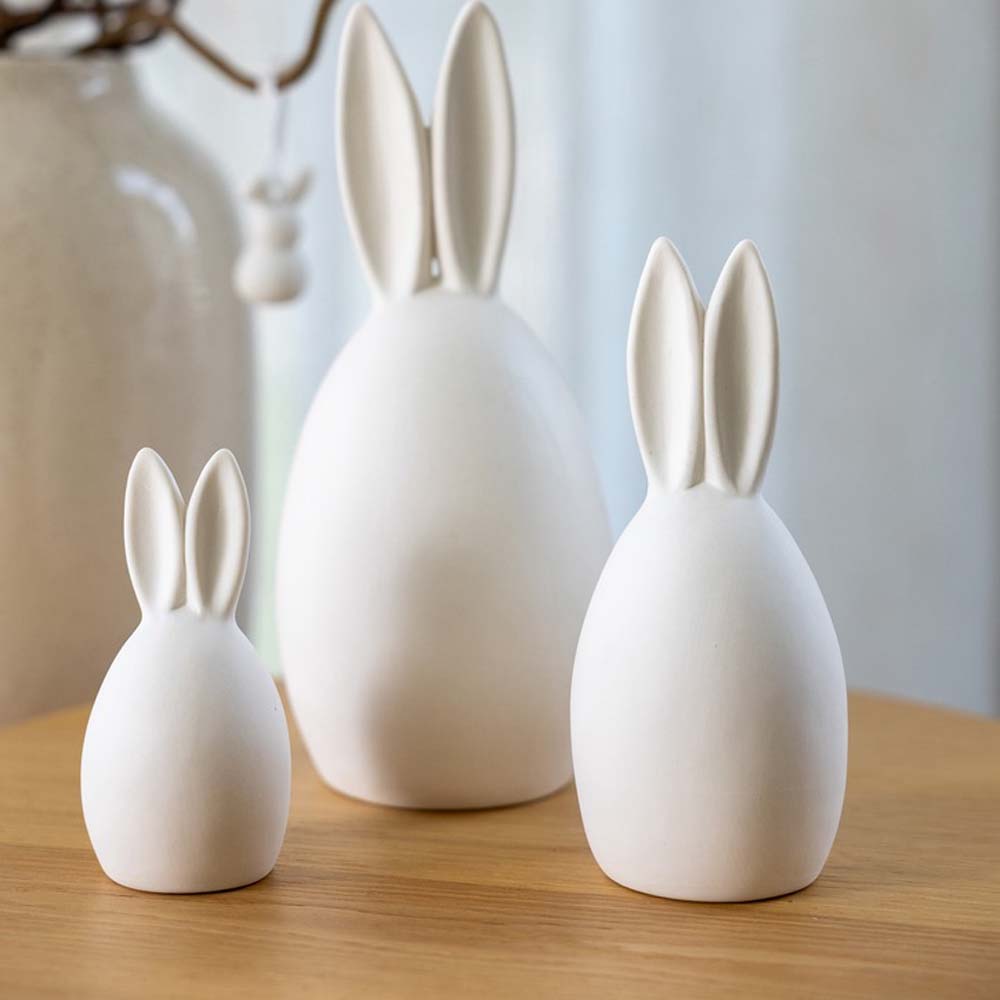 Three Storefactory – Hase Emma, weiße Hasenskulpturen aus Keramik in verschiedenen Größen auf einer Holzoberfläche.