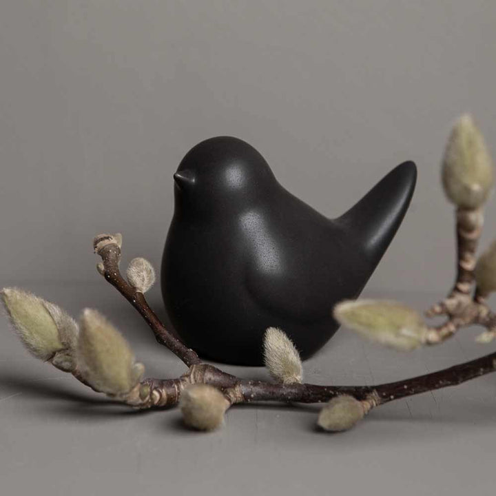 Eine Vogelfigur aus Keramik von Herman Vogel von Storefactory, die auf einem Ast mit knospenden Weidenkätzchen vor einem grauen Hintergrund thront.