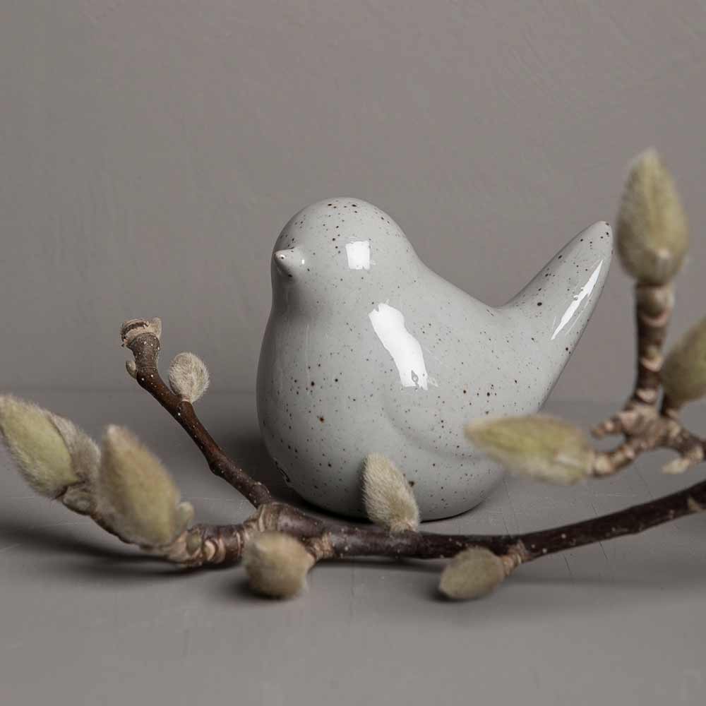 Storefactory – Herman Vogel-Figur in gesprenkeltem Grau, auf einem Ast mit knospenden Weidenkätzchen vor neutralem Hintergrund sitzend.