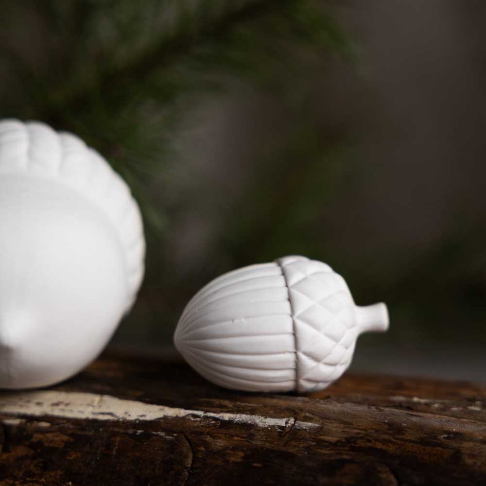 Storefactory – Holeby Eichel, eine weiße Keramik-Eichel, platziert auf einer Holzoberfläche mit Grün im Hintergrund.