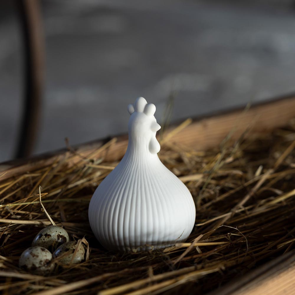 Das Storefactory - Huhn Kerstin ist eine weiße Keramik-Hühnerfigur auf Stroh, begleitet von drei kleinen, gesprenkelten Eiern.