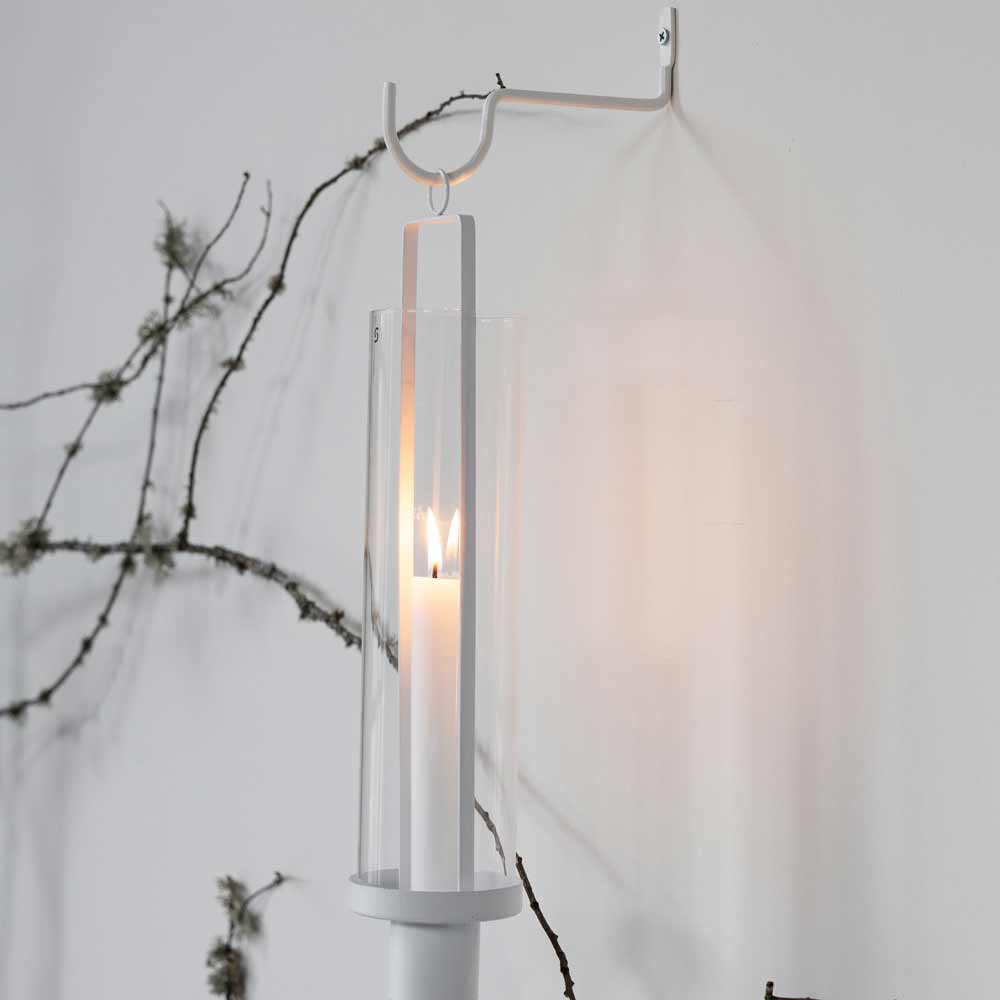 Eine brennende Kerze in einem klaren Glashalter ist mit dem Storefactory - Krokabo Metallhaken Weiß an einer weißen Wand befestigt. Im Hintergrund sind Zweige mit spärlichen Blättern zu sehen.