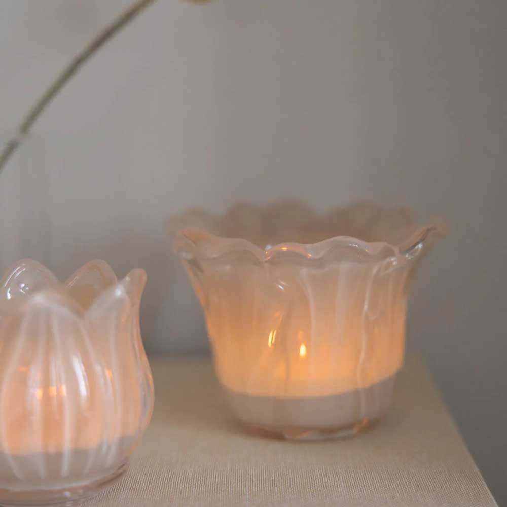 Zwei brennende Storefactory Ljunga Teelichthalter-Kerzen auf einer beigen Oberfläche, mit einem verschwommenen Blütenstiel im Hintergrund.