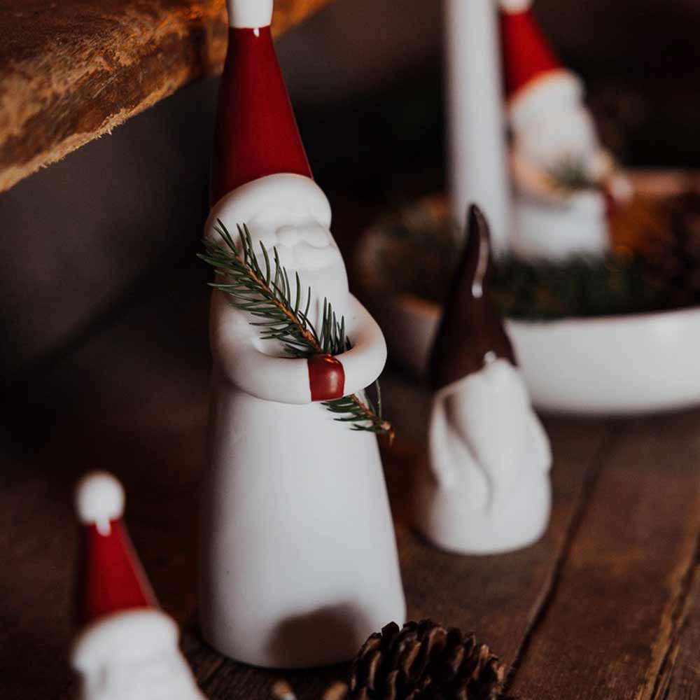 Die Keramikfiguren „Loke Weihnachtsmann aus Keramik“ von Storefactory – Zwerge mit roten Mützen, die Tannenzweige halten, werden auf einer Holzoberfläche präsentiert.