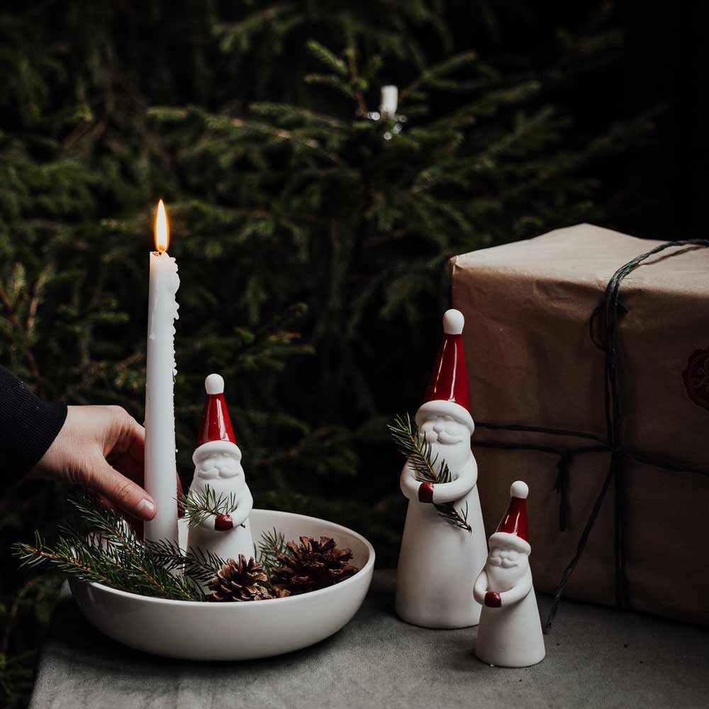 Eine Hand stellt eine brennende weiße Kerze in eine Schale mit Tannenzapfen und Loke-Weihnachtsmann-aus-Keramik-Figuren von Storefactory mit roten Mützen, neben eine in braunes Papier eingewickelte Geschenkbox, im Hintergrund ist ein Weihnachtsbaum zu sehen.