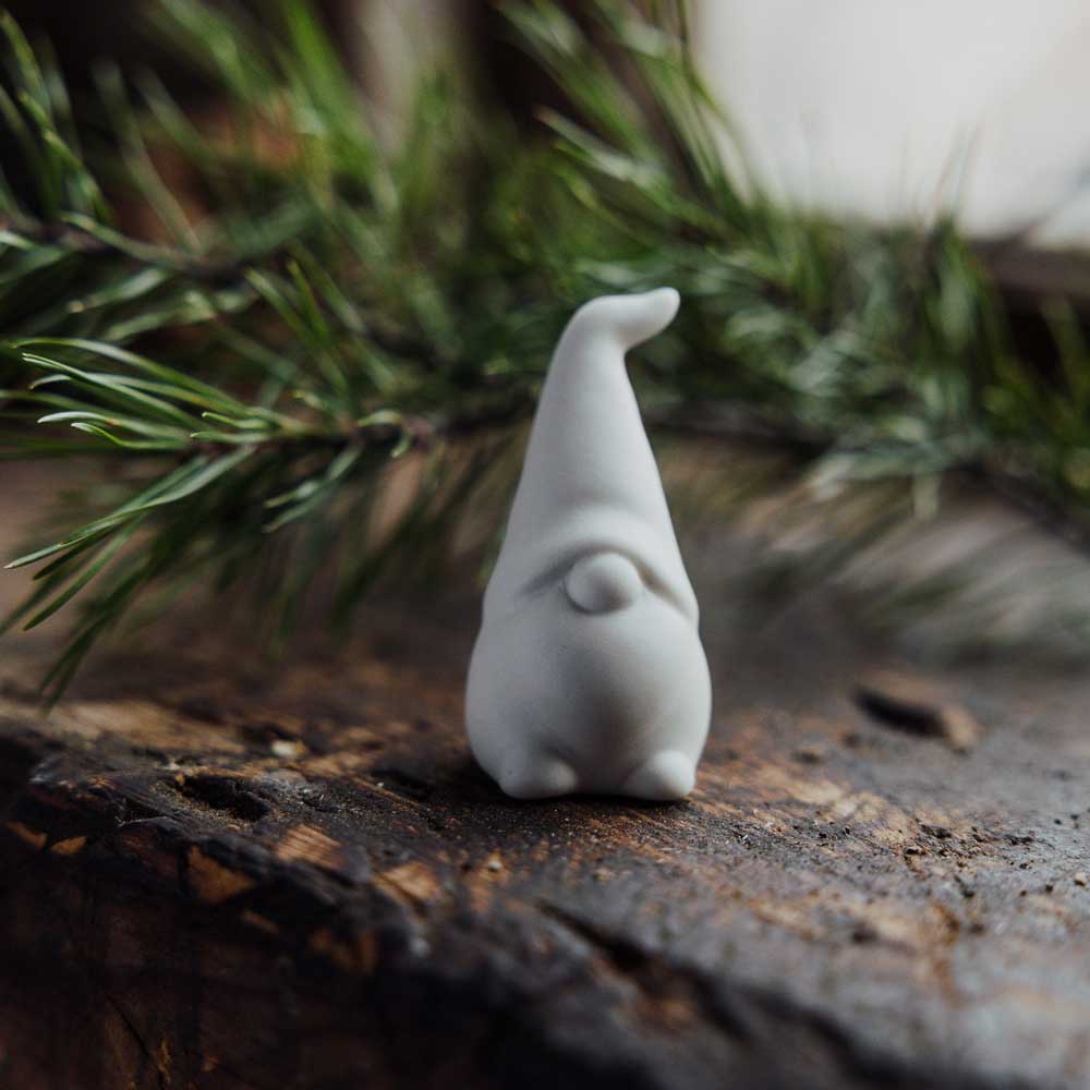 Die Storefactory - Malte Wichtel Keramik weiß, eine kleine Wichtelfigur mit hohem Hut, steht auf einer Holzfläche mit Tannenzweigen im Hintergrund.