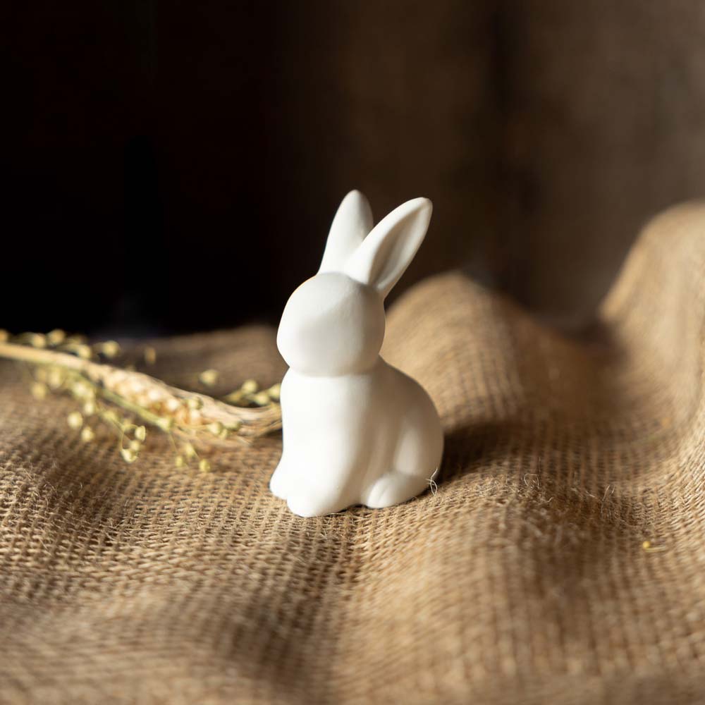 Die Storefactory – Oskar Hase, eine kleine weiße Hasenfigur aus Keramik, ruht auf strukturiertem Sackleinen mit trockenem Gras im Hintergrund.