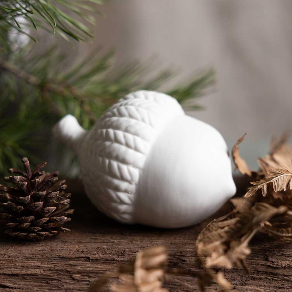 Västansjö Keramik Eichel von Storefactory auf einer Holzoberfläche platziert, umgeben von Tannenzweigen, Tannenzapfen und getrockneten Blättern.