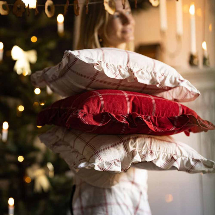 Eine Person hält einen Stapel Strömshaga - Kissenbezug Alma 45 x 45 cm Kissen mit einem verschwommenen Weihnachtsbaum im Hintergrund.