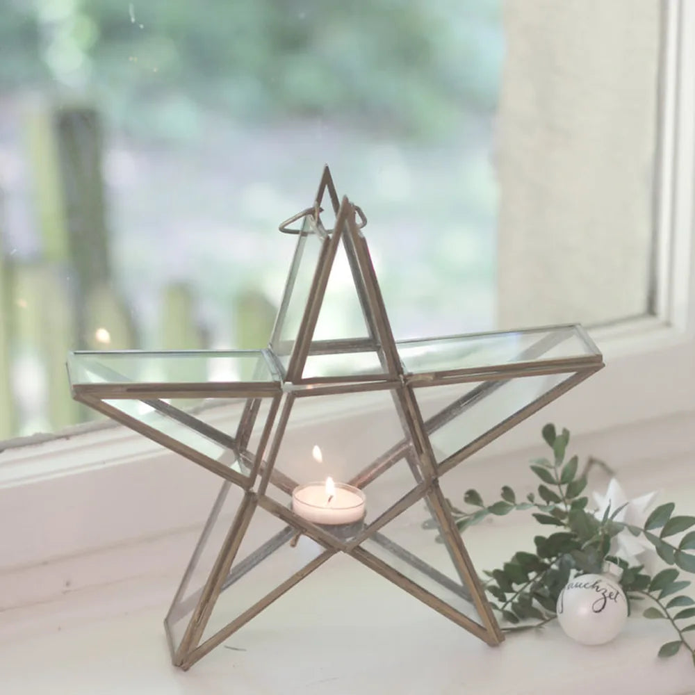 Der VL Home Glasstern für Teelicht zum Hängen und Stellen, mit einem brennenden Teelicht, steht auf der Fensterbank neben einer kleinen Dekofigur und etwas Grün.