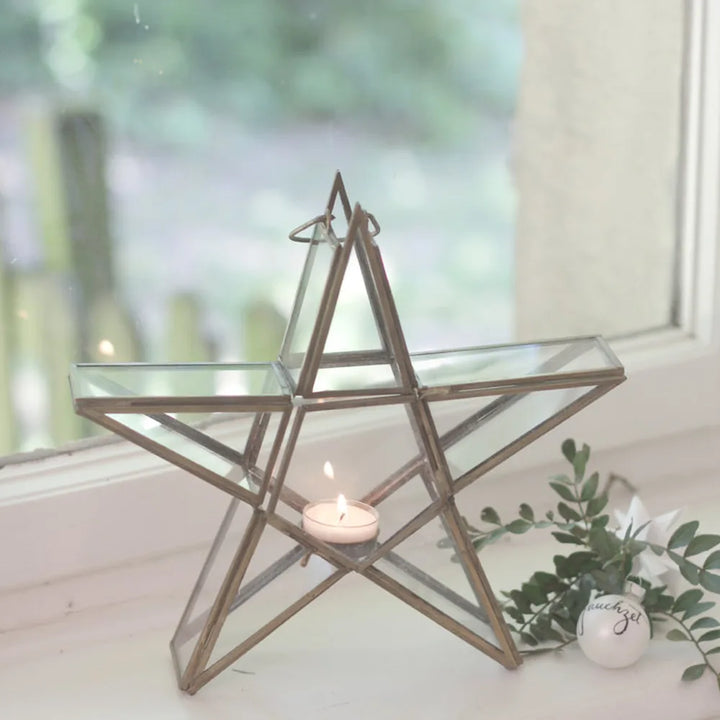 Der VL Home Glasstern für Teelicht zum Hängen und Stellen, mit einem brennenden Teelicht, steht auf der Fensterbank neben einer kleinen Dekofigur und etwas Grün.