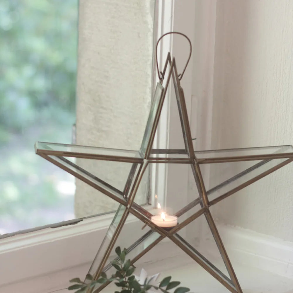 Der VL Home Glasstern für Teelicht zum Hängen und Stellen aus Glas und Metall steht mit einem brennenden Teelicht auf der Fensterbank, während im Vordergrund grünes Laub zu sehen ist.