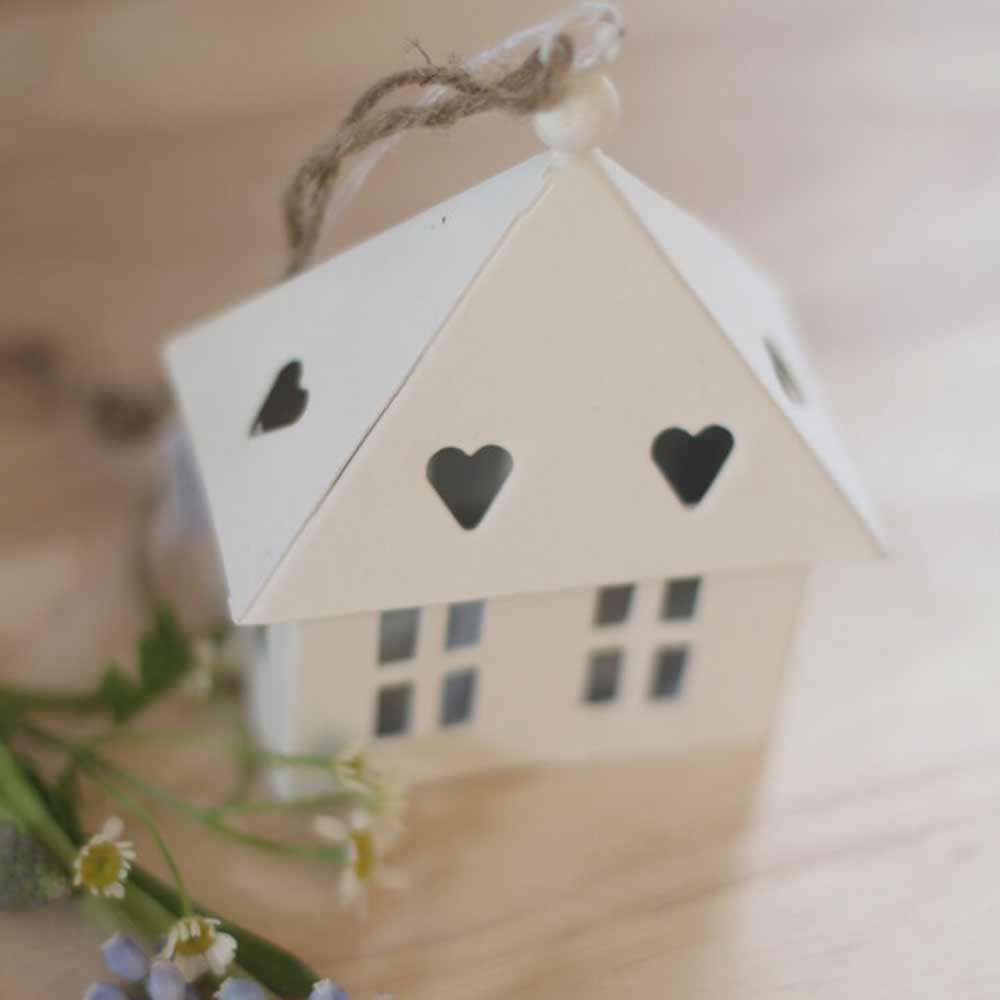 Das VL-Home - Haus rechteckig zum Hängen mit Herzen im Dach ist ein kleines weißes Deko-Haus mit herzförmigen Ausschnitten und einer Schnurschlaufe, das auf einer Holzfläche neben Blumen präsentiert wird.