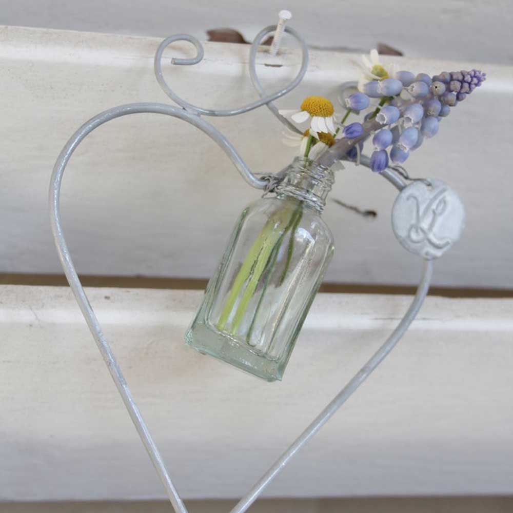 Clear VL Home – Herz mit 1 Miniflasche mit Blumen, darunter violette und gelbe Blüten, in einem herzförmigen Halter aus Metalldraht vor einem weißen Holzhintergrund.