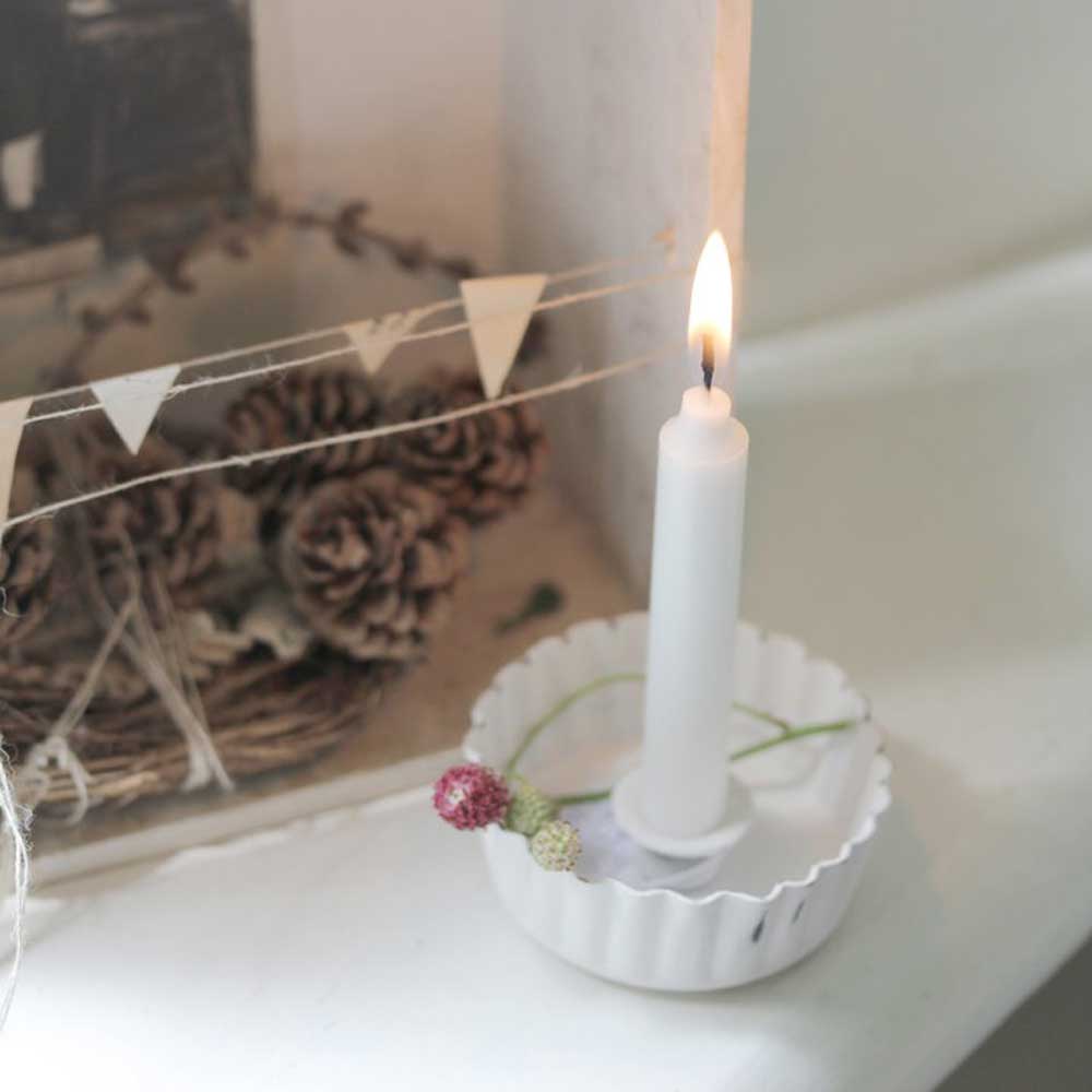 Ein VL Home - Kerzenhalter Backform weiß brennt in einem gewellten Halter mit Blumendekor, neben einem geflochtenen Korb mit Tannenzapfen und einer Schnur mit kleinen Fähnchen platziert.