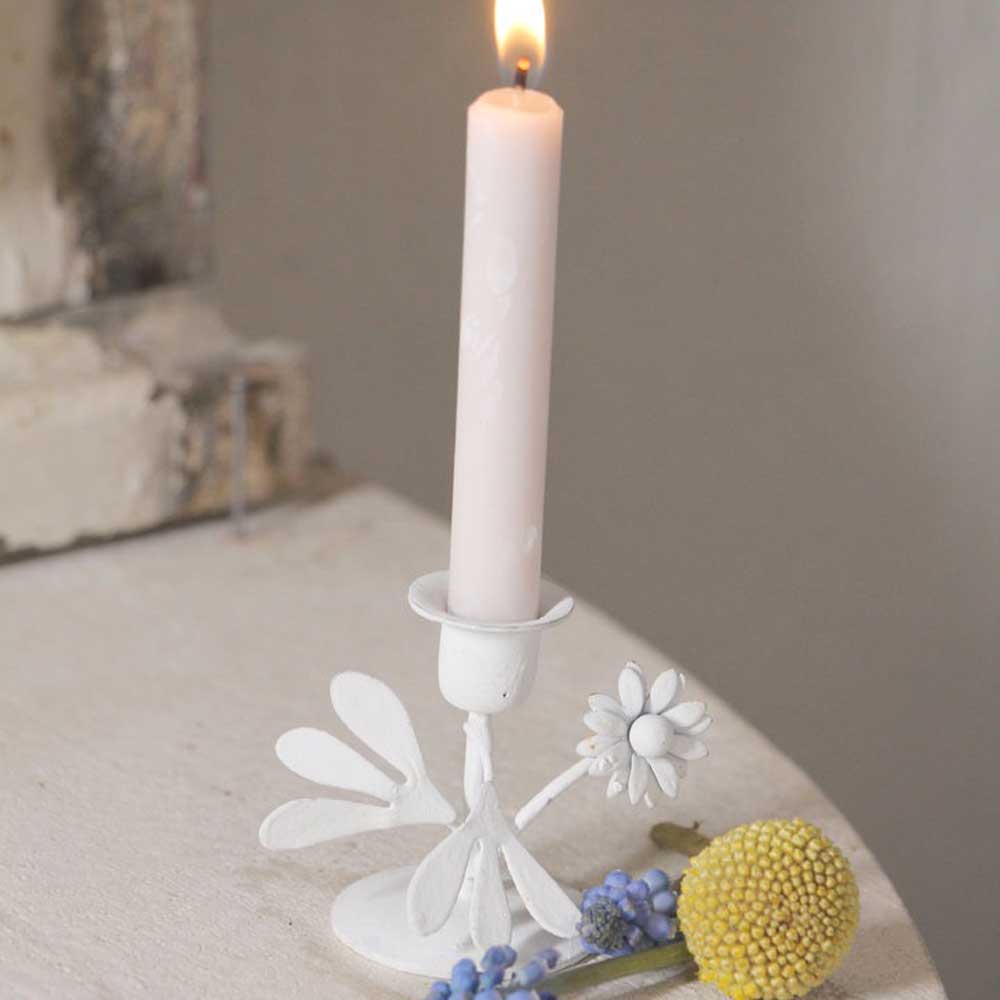 Eine brennende rosa Kerze wird von einem weißen VL Home - Kerzenhalter Blumen in Form einer Person mit Blumen gehalten, neben einer Gruppe gelber und blauer Blumen auf einer weißen Oberfläche.