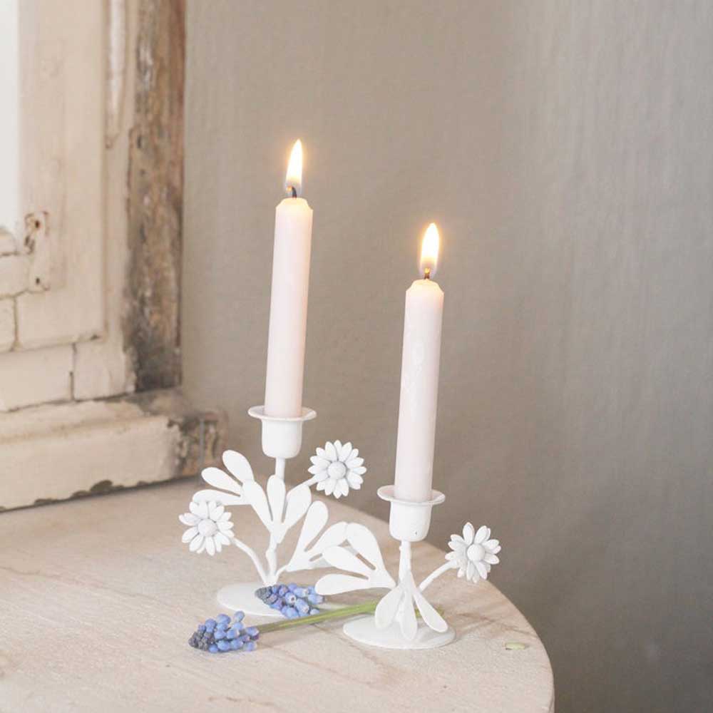 Zwei brennende weiße Kerzen in einem VL Home - Kerzenhalter Blumen, platziert auf einer hellen Oberfläche neben einem kleinen Strauß blauer Blumen. Im Hintergrund ist ein verwitterter Fensterrahmen zu sehen.