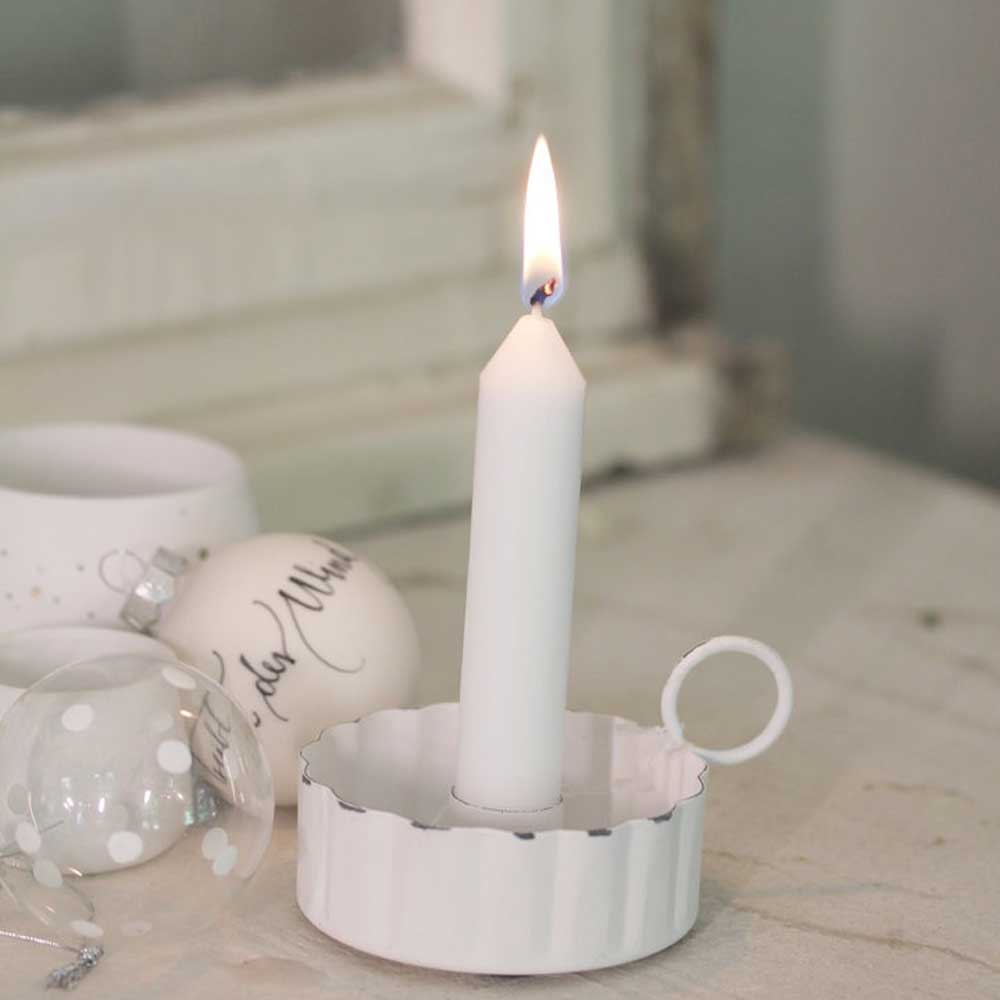 Eine brennende weiße Kerze in einem VL Home – Kerzenhalter mit Griff für Stabkerzen gewellter Rand, platziert auf einer hellen Oberfläche mit weißen Ornamenten im Hintergrund.