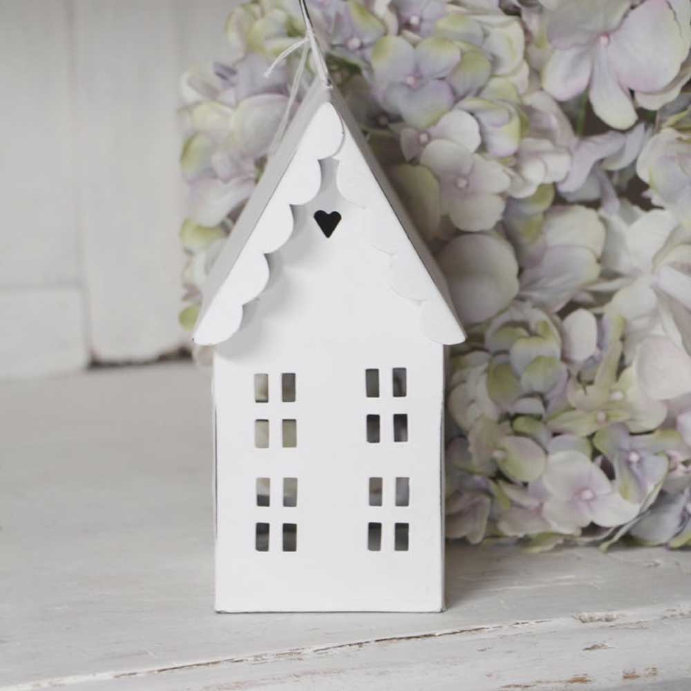 Ein kleines, weißes, dekoratives VL Home - Metallhaus Deko weiß Herz im Dach 2 Etagen mit gewellten Kanten und einem Herzausschnitt auf dem Dach steht vor hellen Hortensienblüten.