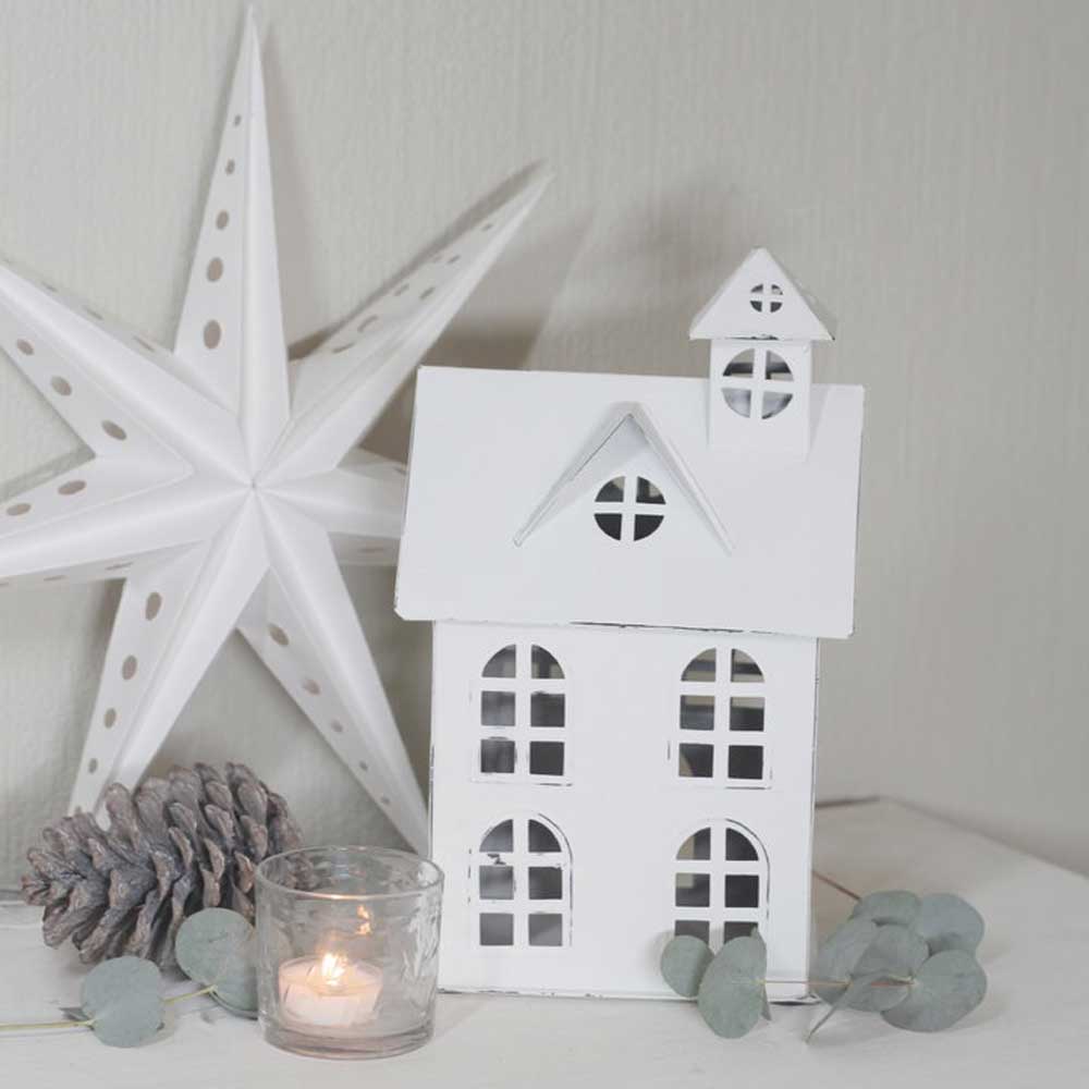 Ein VL Home – Metallhaus mit Schornstein mit einem Sternornament, einem Tannenzapfen und einer brennenden Kerze in einem Glashalter auf hellem Hintergrund.