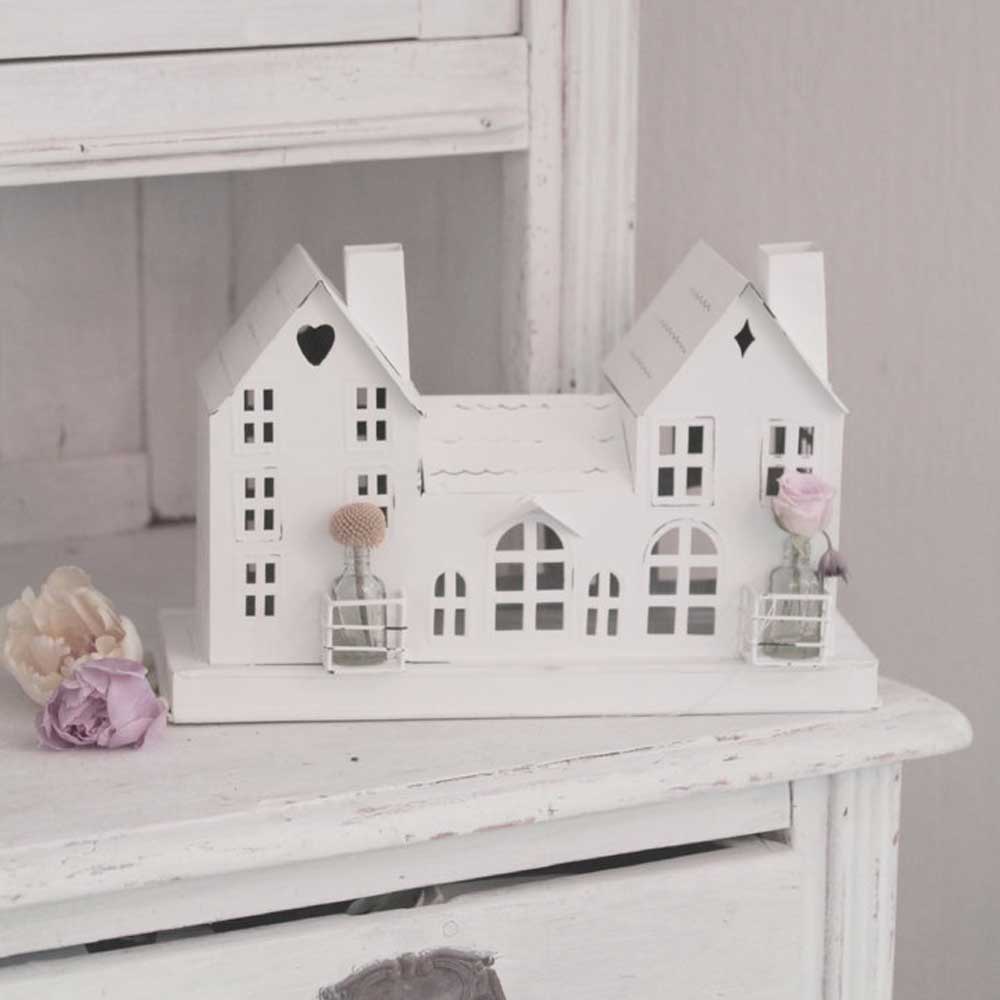 Auf einer weißen Holzoberfläche steht ein dekoratives weißes Haus von VL Home - Metallhaus weiß, Häuserreihe Deko mit 3 Häusern mit kleinen Glasgefäßen voller Blumen.