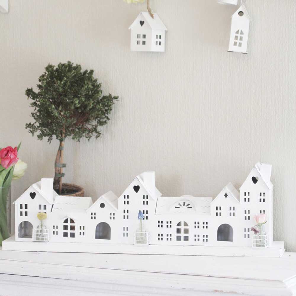Ein dekoratives Display „VL Home - Metallhaus weiß Häuserreihe Deko mit 7 Häusern“ steht auf einem Regal mit kleinen Vasen voller Blumen. Im Hintergrund sind ein kleiner Baum und hängende Hausornamente zu sehen.