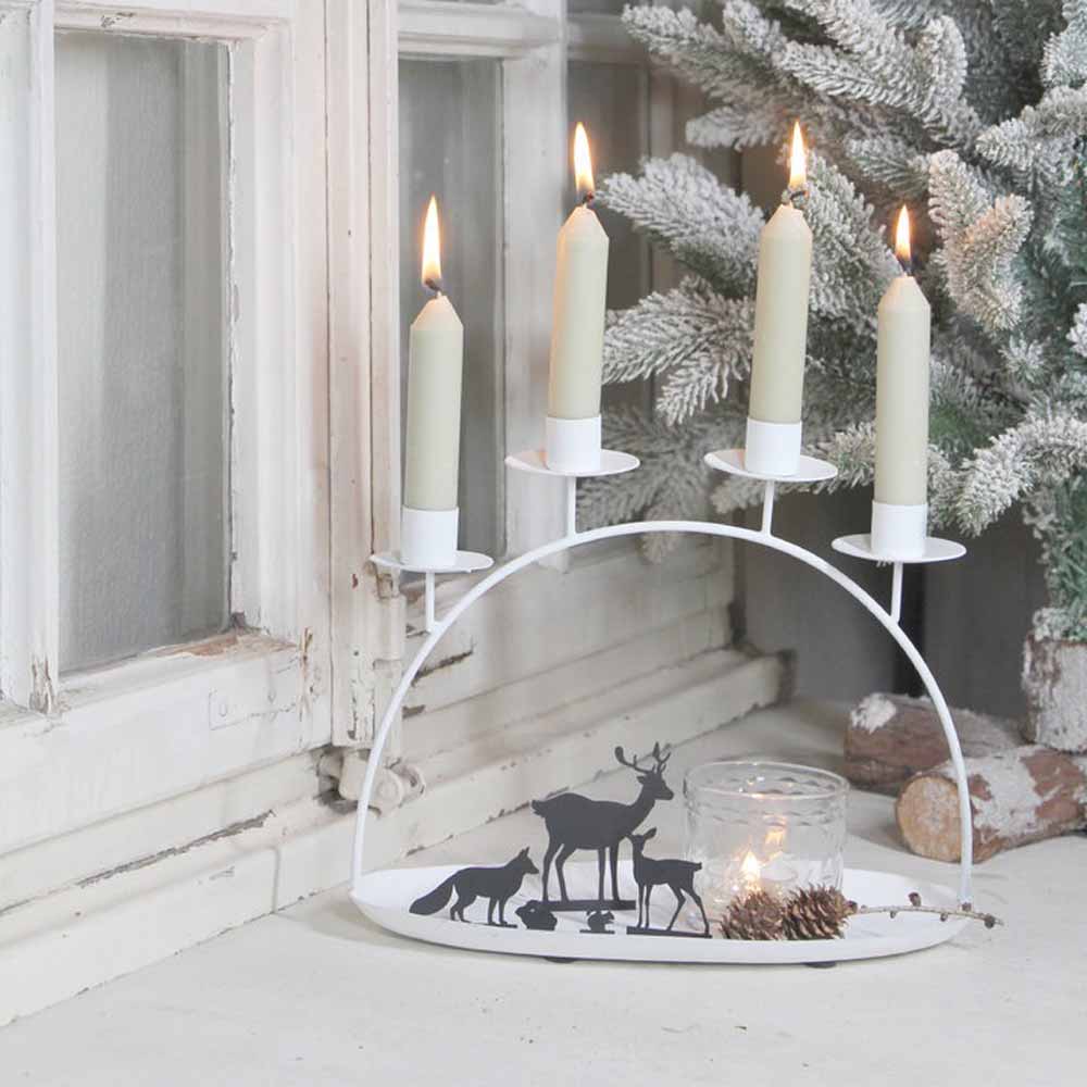 Die VL Home Schale mit Kerzenbogen für vier Stabkerzen ist beleuchtet und zeigt Tiersilhouetten auf einem weißen bogenförmigen Halter, der neben einem Milchglasfenster und einem schneebedeckten künstlichen Baum platziert ist.
