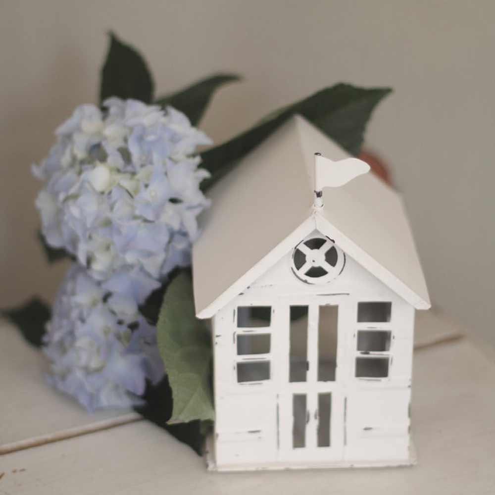 Das VL Home – Strandhaus Metallhaus Tür mit Fenster, ein kleines weißes Dekohaus, steht neben hellblauen Hortensienblüten und grünen Blättern.
