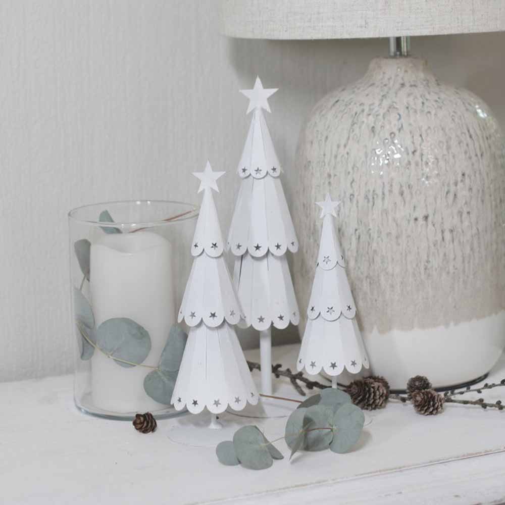 Der VL Home – Tannenbaum Metall mit Sternen und ein mit Eukalyptusblättern geschmückter Kerzenhalter aus Glas sind auf einer weißen Oberfläche neben einer strukturierten Lampe arrangiert.