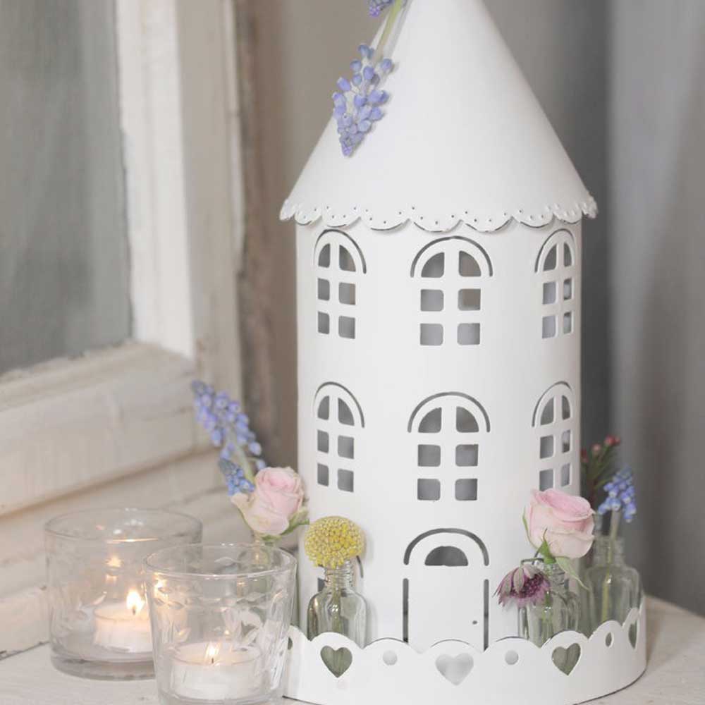 Eine weiße, hausförmige Kerzenlaterne mit Blumendekorationen und drei kleinen Kerzenhaltern aus Glas, in denen jeweils eine Kerze brennt, steht auf einer weißen Fläche neben einem Fenster. VL Home – Vogelhaus rund mit Glasflaschen kommt in dieser ruhigen Umgebung wunderbar zur Geltung.